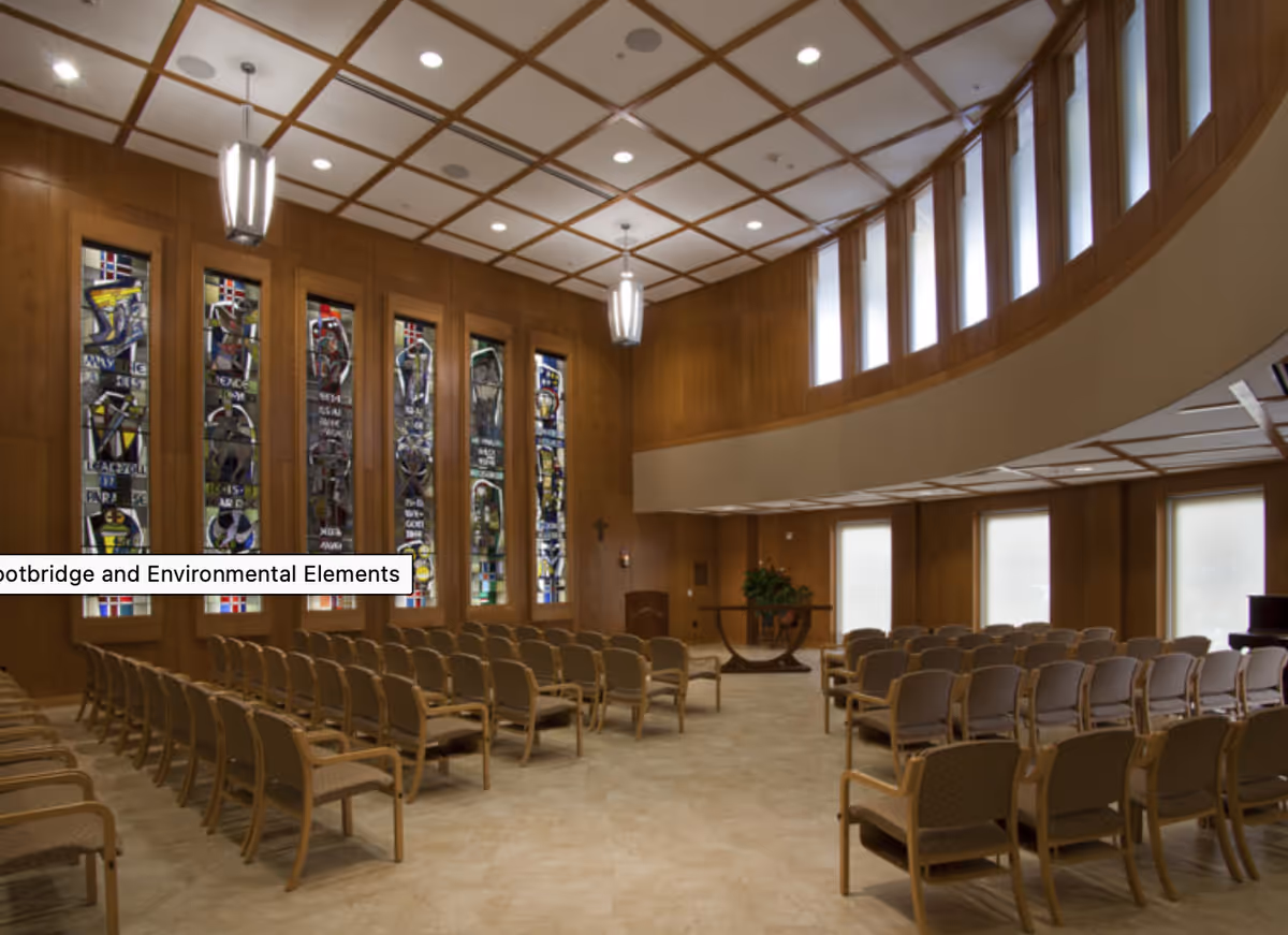 Interior view of a chapel or worship room with rows of beige cushioned chairs arranged facing a wooden podium. The room features tall stained glass windows on one wall and a curved upper section with vertical windows letting in natural light. The ceiling has a grid pattern with recessed lighting and hanging light fixtures.
