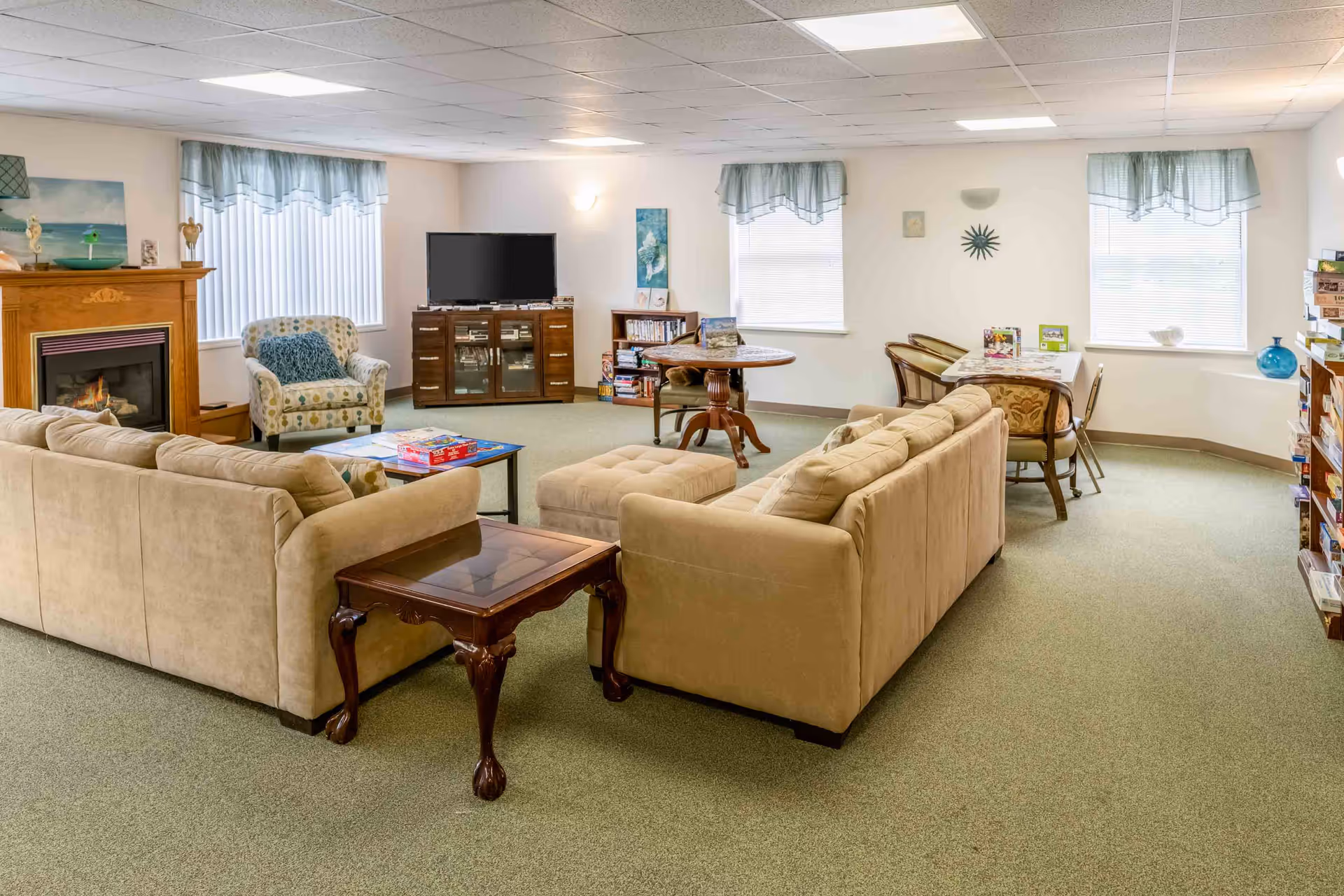 A spacious living room area with beige sofas arranged around a coffee table with board games on it. There is a fireplace with a wooden mantel on the left, an armchair with a blue cushion, and a TV on a wooden stand against the wall. The room has several windows with light blue valances, a round wooden table, and a rectangular table with chairs. Shelves with board games and books are visible along the walls.