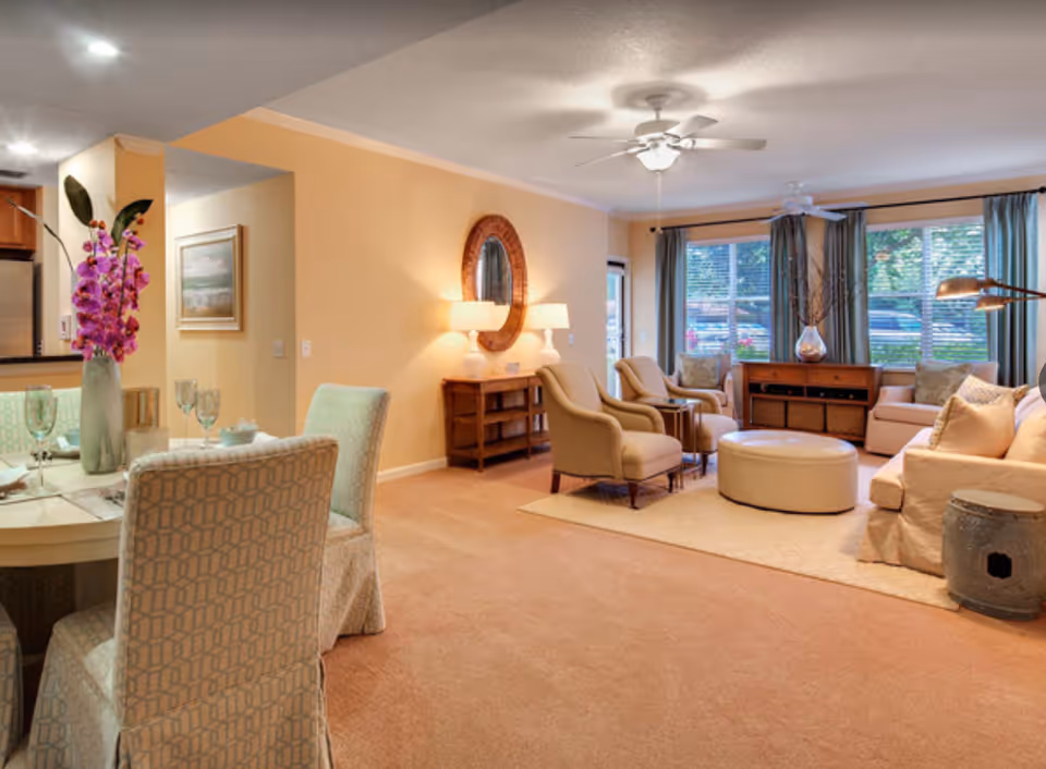 A cozy living room and dining area in a senior living facility with beige walls and carpet. The living room features a beige sofa, two armchairs, a round ottoman, a wooden console table with a vase, and large windows with blue curtains letting in natural light. The dining area has a round table set with glassware and a tall vase with purple flowers. A ceiling fan with a light fixture is mounted on the ceiling.