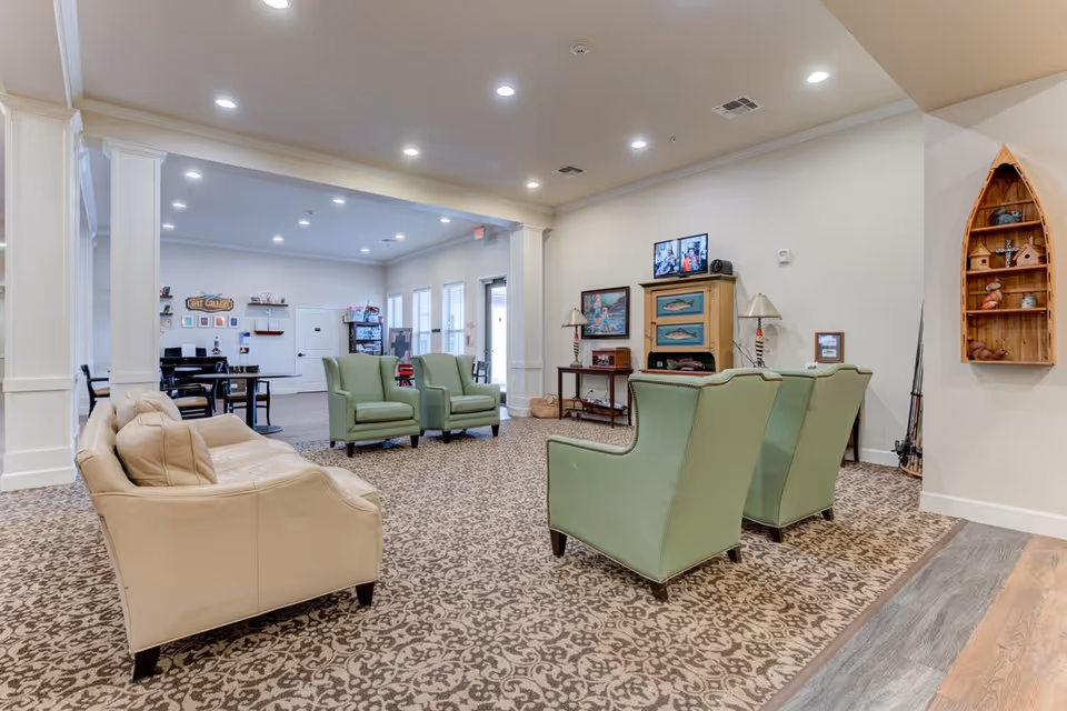 A senior living facility common area with beige and green armchairs arranged on a patterned carpet. The room has white walls and ceiling with recessed lighting. There is a TV mounted on the wall above a cabinet decorated with fish artwork. A wooden boat-shaped shelf with decorative items is mounted on the right wall. In the background, there is a dining area with tables and chairs near windows.