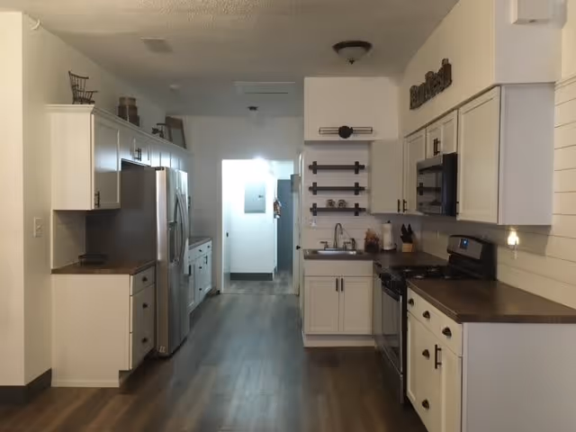 A modern kitchen with white cabinets, stainless steel refrigerator, stove, and microwave. The kitchen has wooden countertops and dark wood flooring. There are decorative items on the shelves and counters, and a hallway is visible in the background.
