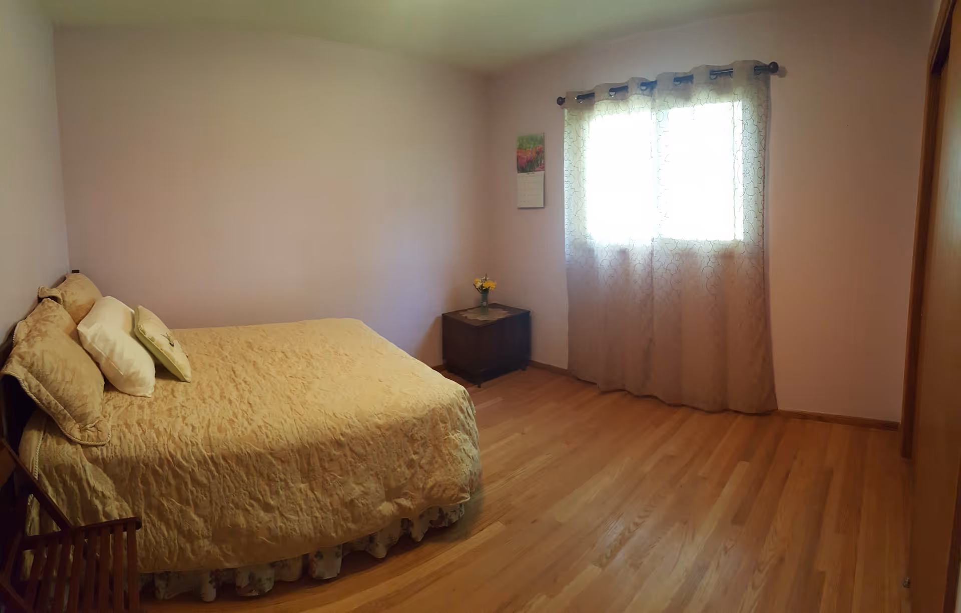 A simple bedroom with a bed covered in a beige quilt and several pillows. There is a small wooden nightstand with a vase of flowers next to the bed. A window with sheer curtains allows light into the room, and a calendar hangs on the wall beside the window. The floor is wooden, and the walls are painted a light color.