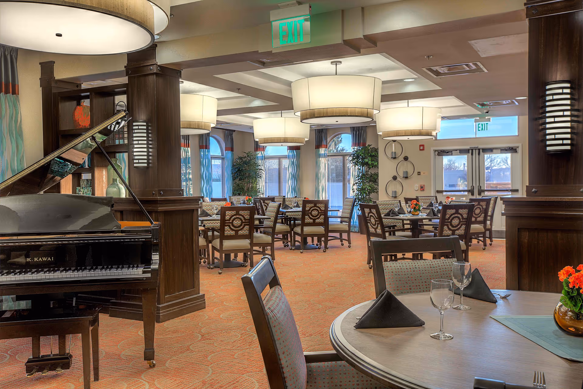A spacious and well-lit dining room in an assisted living facility featuring round tables set with black napkins, wine glasses, and floral centerpieces. The room has large windows with curtains, modern ceiling lights, and a black grand piano on the left side. The decor includes wooden pillars and warm-toned carpeting.