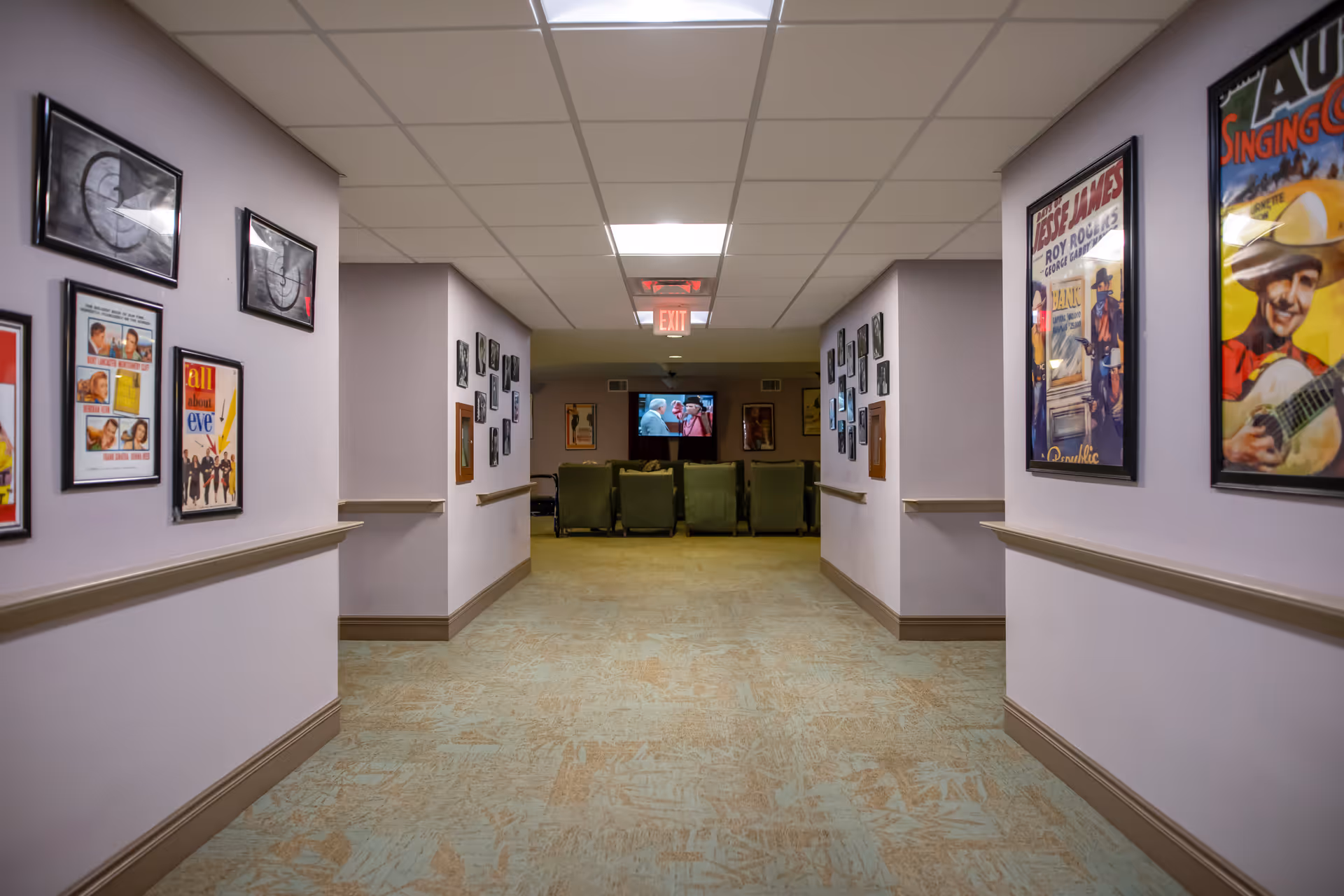 A softly lit hallway in a senior living facility lined with framed posters leading to a small seating area with a TV.