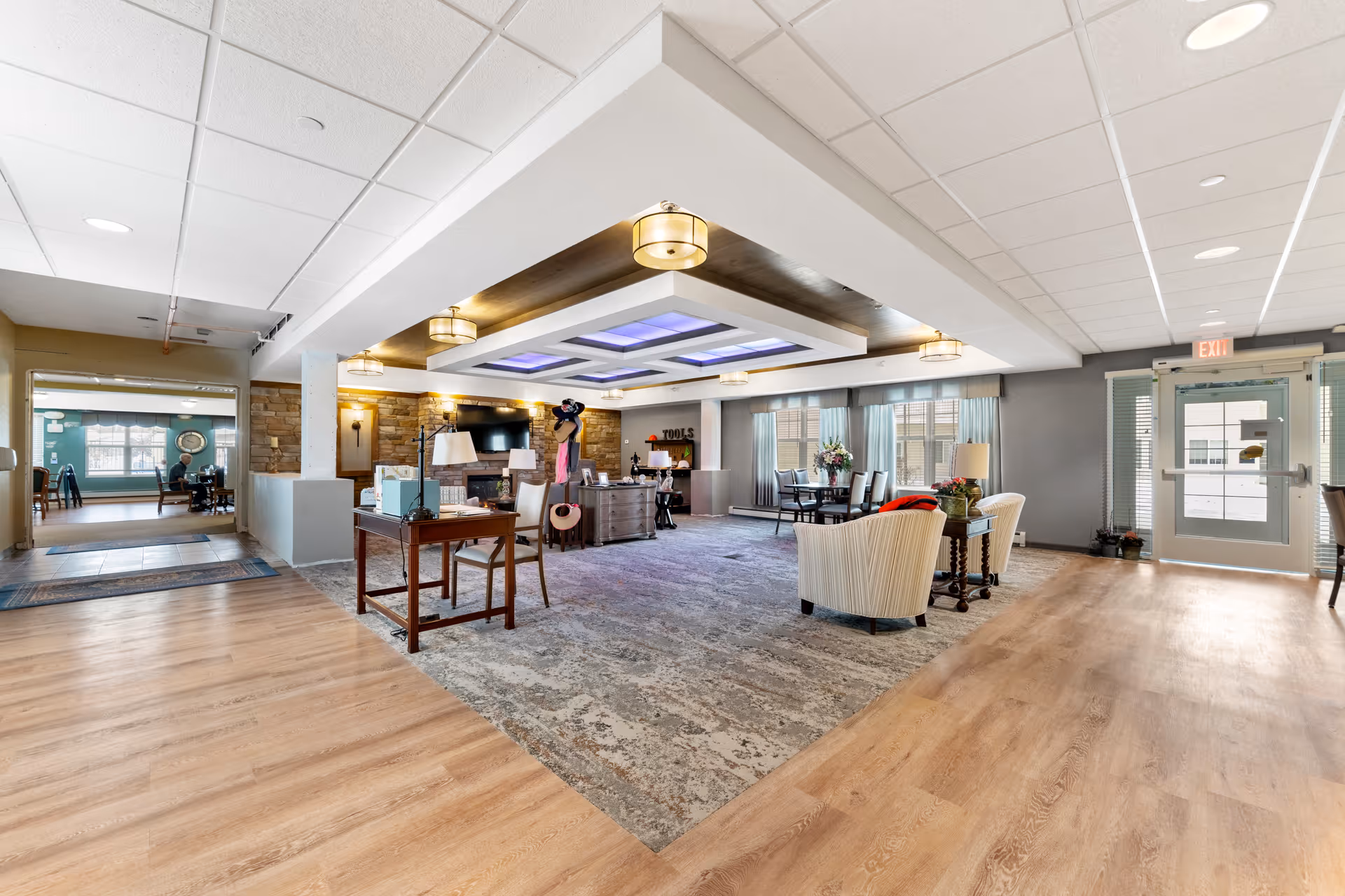 A spacious senior living facility common area with a mix of wooden and carpeted flooring. The room features comfortable armchairs, a table with chairs near windows with curtains, a desk with a chair, and a decorative ceiling with recessed lighting. There is a door with glass panels on the right side and an adjacent room visible through an open doorway on the left.