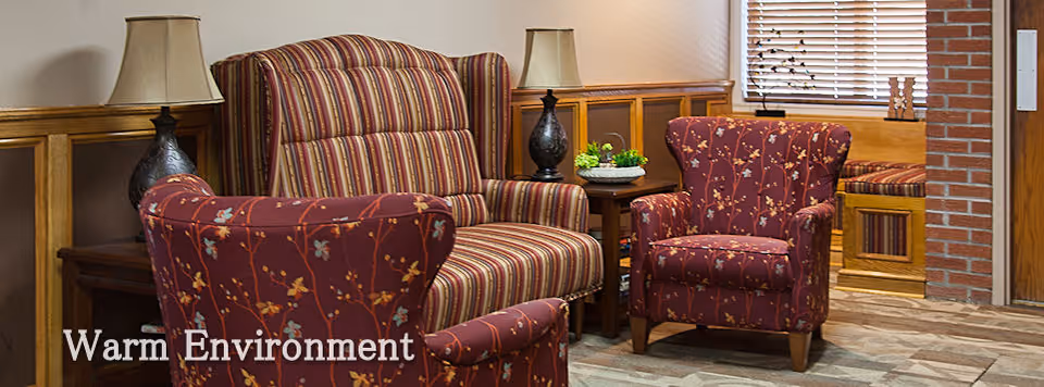 A cozy seating area with two floral patterned armchairs and a striped loveseat, two table lamps on wooden side tables, a small plant on one table, wood paneling on the walls, a window with blinds, and a brick column near the entrance door.