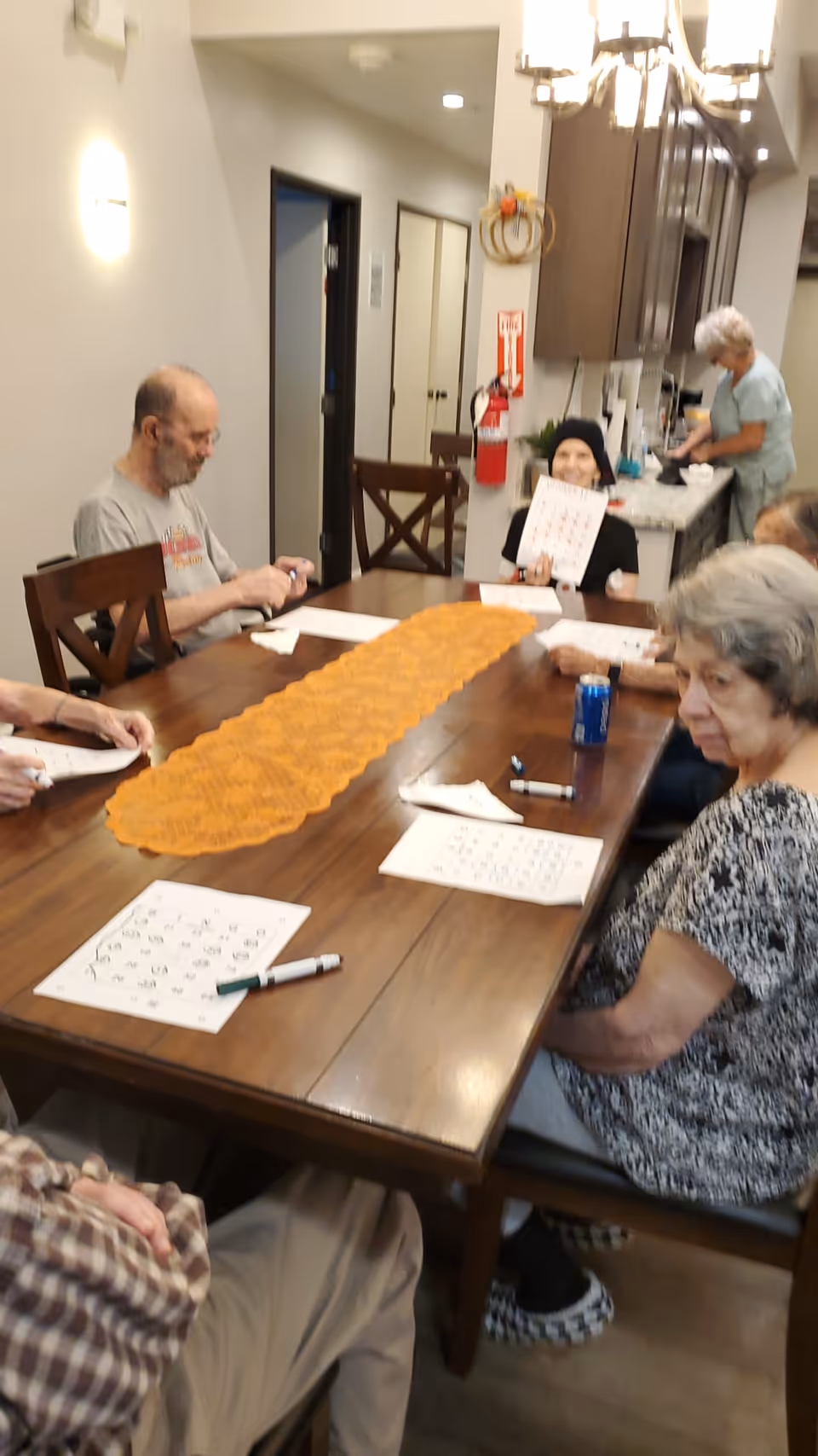 A group of elderly people sitting around a wooden dining table playing bingo. One person is holding up a bingo card and smiling. There are bingo cards and markers on the table, and a woman is standing in the kitchen area in the background. The room has warm lighting and a cozy atmosphere.