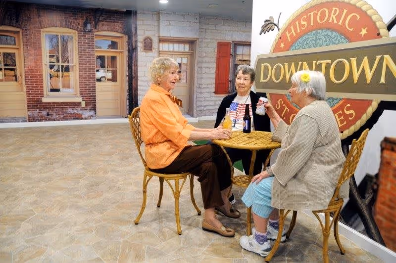Three elderly women sitting around a small round wicker table with drinks, engaged in conversation inside a room with a large wall mural depicting a historic downtown street scene.
