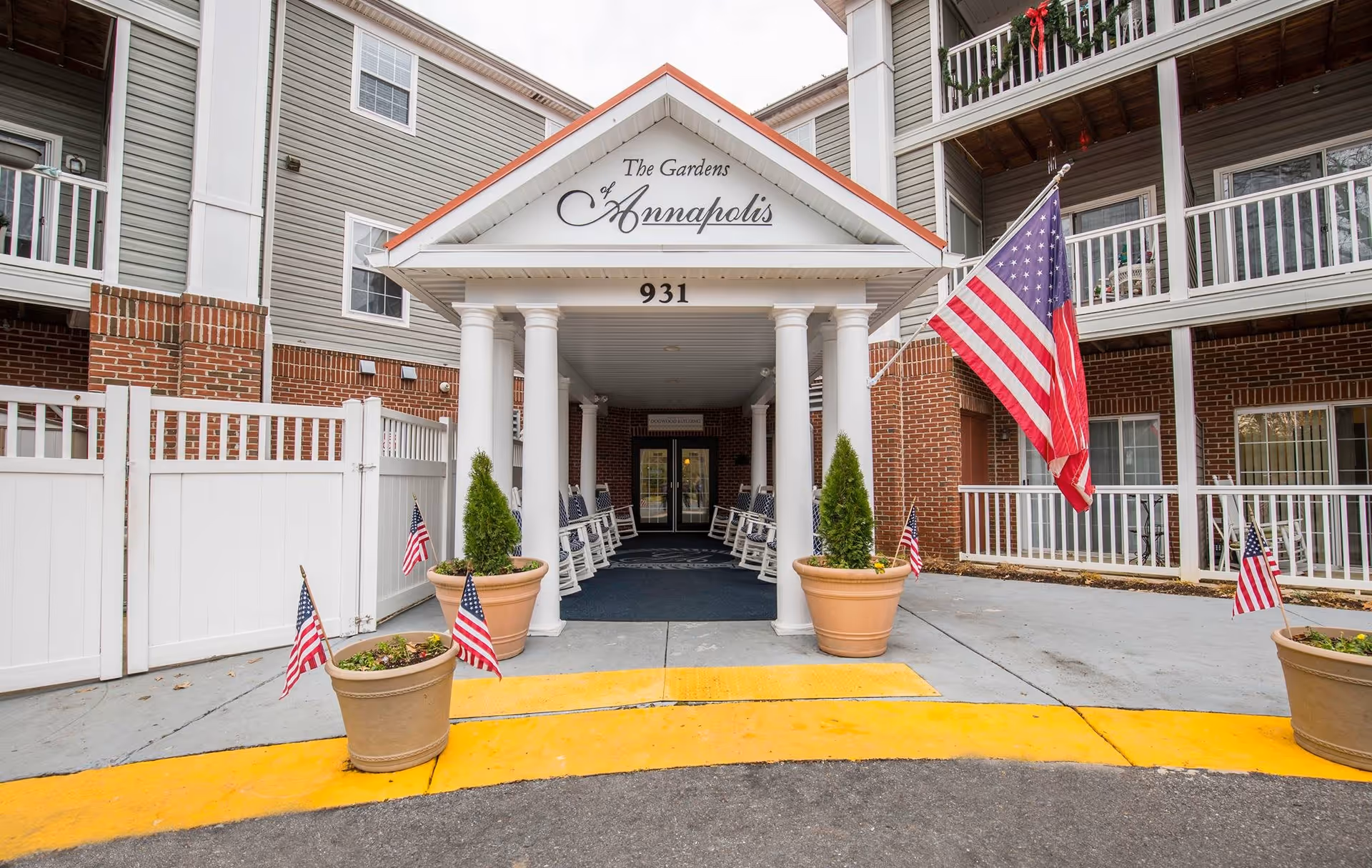Entrance of The Gardens of Annapolis senior living facility with a covered porch supported by white columns, potted plants with American flags, rocking chairs along the walkway, and the building's name displayed above the entrance.
