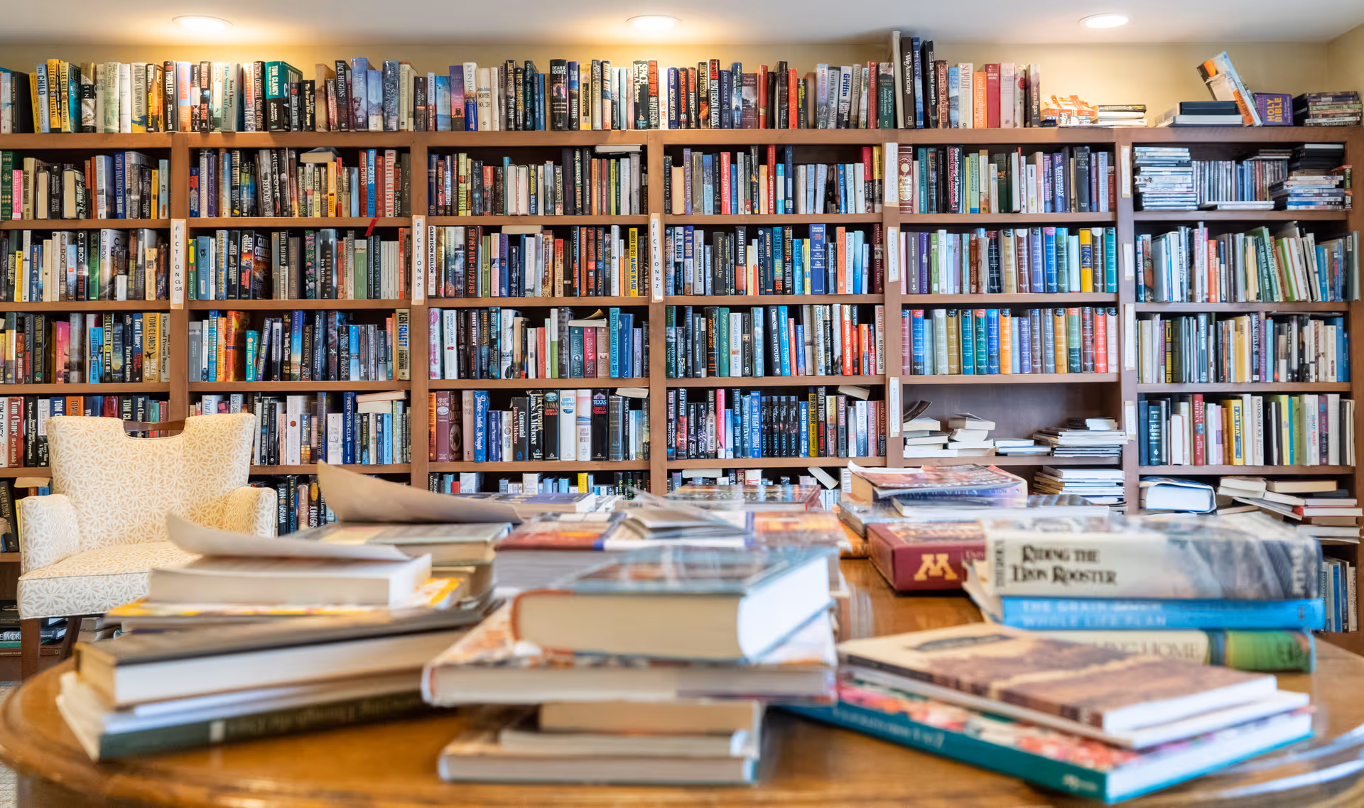 A cozy library room with wooden bookshelves filled with numerous books. In the foreground, there is a wooden table stacked with various books and magazines. To the left, there is a comfortable patterned armchair. The room is warmly lit with ceiling lights.
