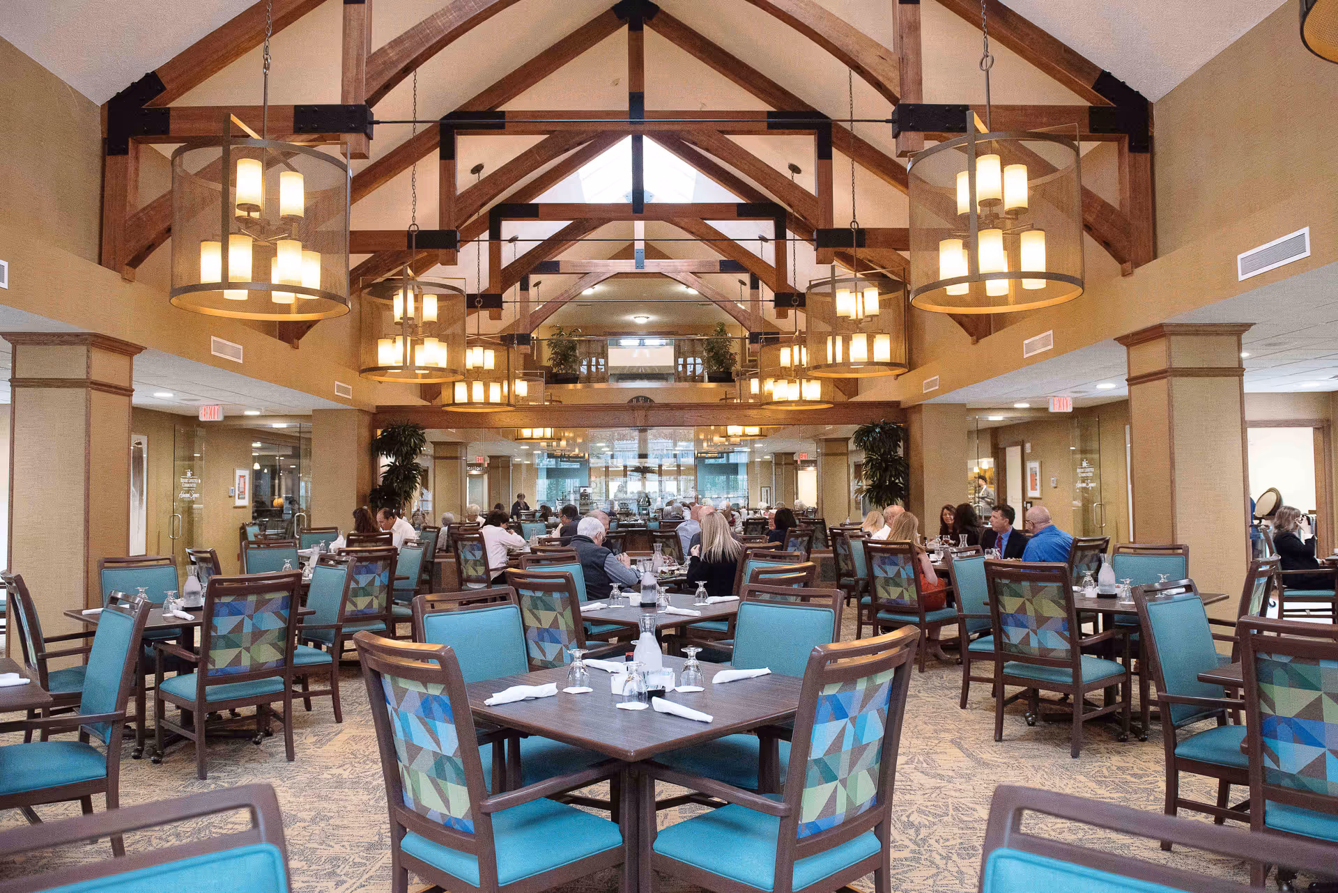 A spacious dining room in a retirement community with multiple tables and chairs arranged neatly. The chairs have blue cushions and patterned backs. Several groups of people are seated and engaged in conversation. The room features high vaulted ceilings with exposed wooden beams and multiple hanging light fixtures providing warm lighting.