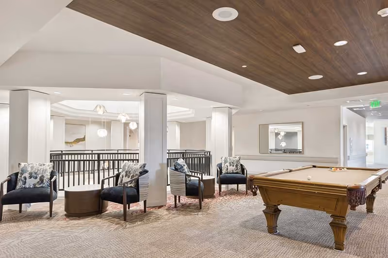 A spacious indoor lounge area featuring a pool table on the right and four cushioned armchairs with floral pillows arranged around two small round tables on the left. The ceiling has wood paneling with recessed lighting, and a large mirror hangs on the wall behind the pool table. The area overlooks a lower floor with a black railing.