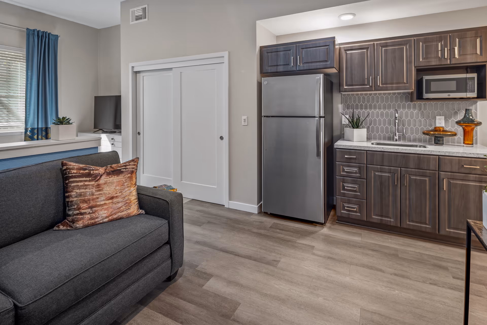 Apartment-style interior with a gray sofa in the foreground and an adjoining kitchenette featuring a stainless refrigerator and dark wood cabinets.