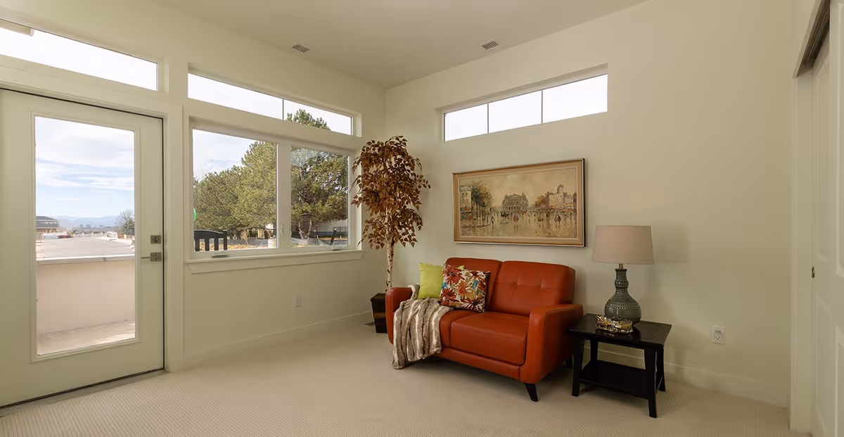 A bright living room with a large window and glass door leading outside. The room features a red leather loveseat with colorful pillows and a throw blanket, a side table with a decorative lamp, a framed painting on the wall, and a tall potted plant in the corner.
