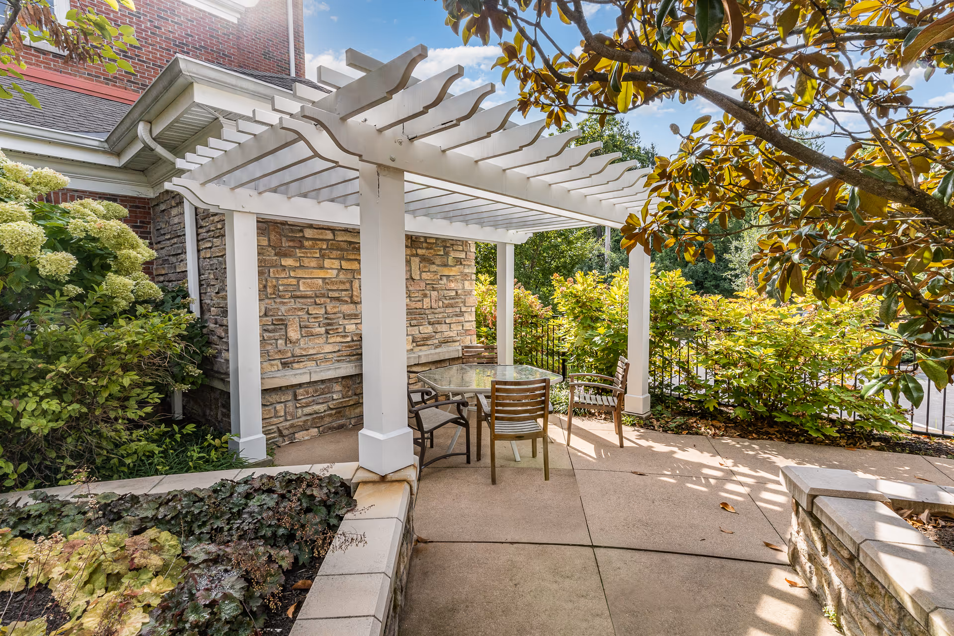 Outdoor patio area with a white pergola providing partial shade over a glass-top table and four wooden chairs. The patio is surrounded by lush greenery, including bushes and trees, with a brick and stone building wall in the background under a clear blue sky.