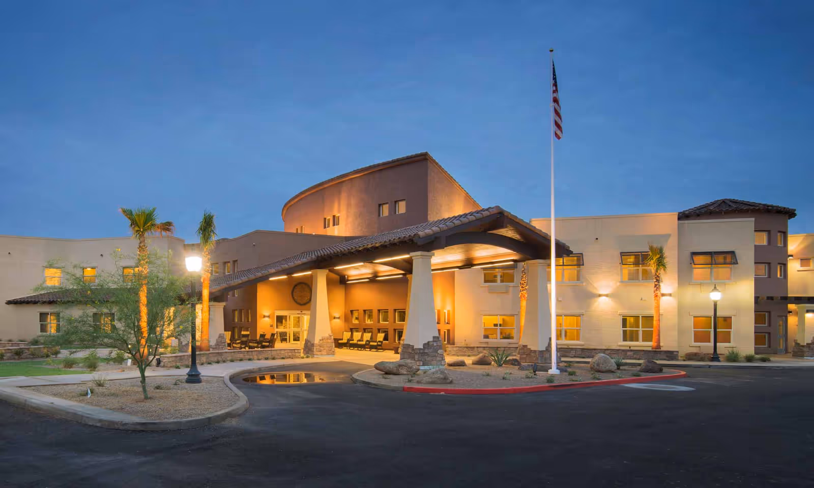 Exterior view of Solana at The Park Assisted Living Community building at dusk, showing a well-lit entrance with a covered driveway, palm trees, an American flag on a tall flagpole, and a clear sky.
