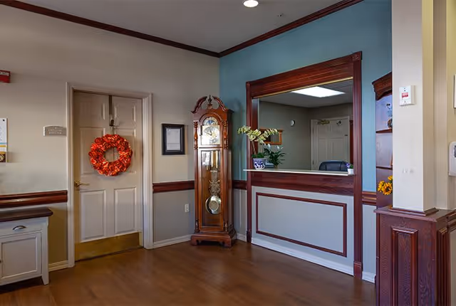 Interior view of a reception area in an assisted living facility featuring a wooden grandfather clock, a door decorated with a red wreath, a reception window with a wooden frame, and a small counter with a potted plant.