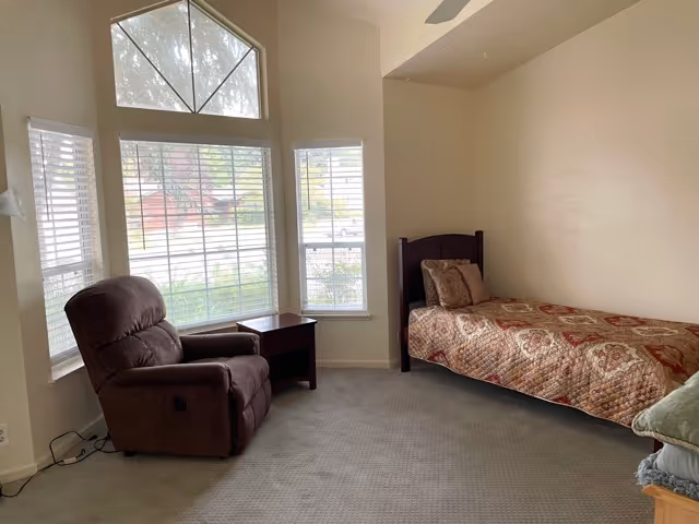 A bright bedroom with a single bed, recliner chair and side table beside large windows.