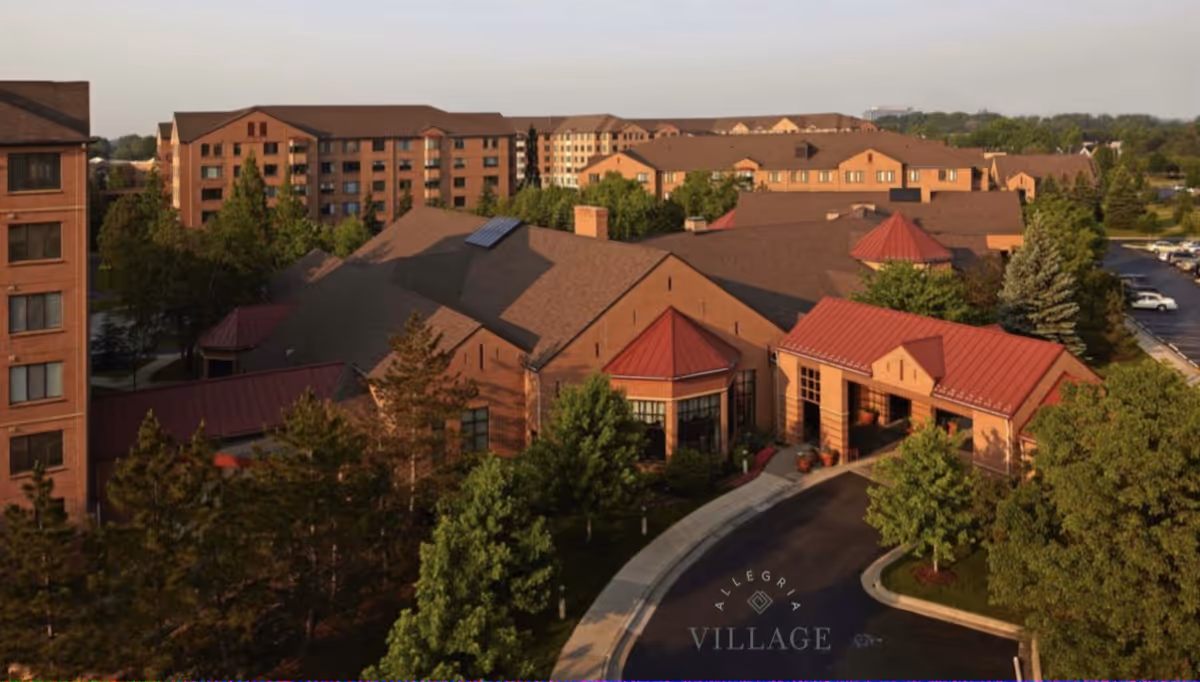 Aerial view of Allegria Village senior living facility showing multiple connected brick buildings with red roofs surrounded by trees and a parking lot in the background.
