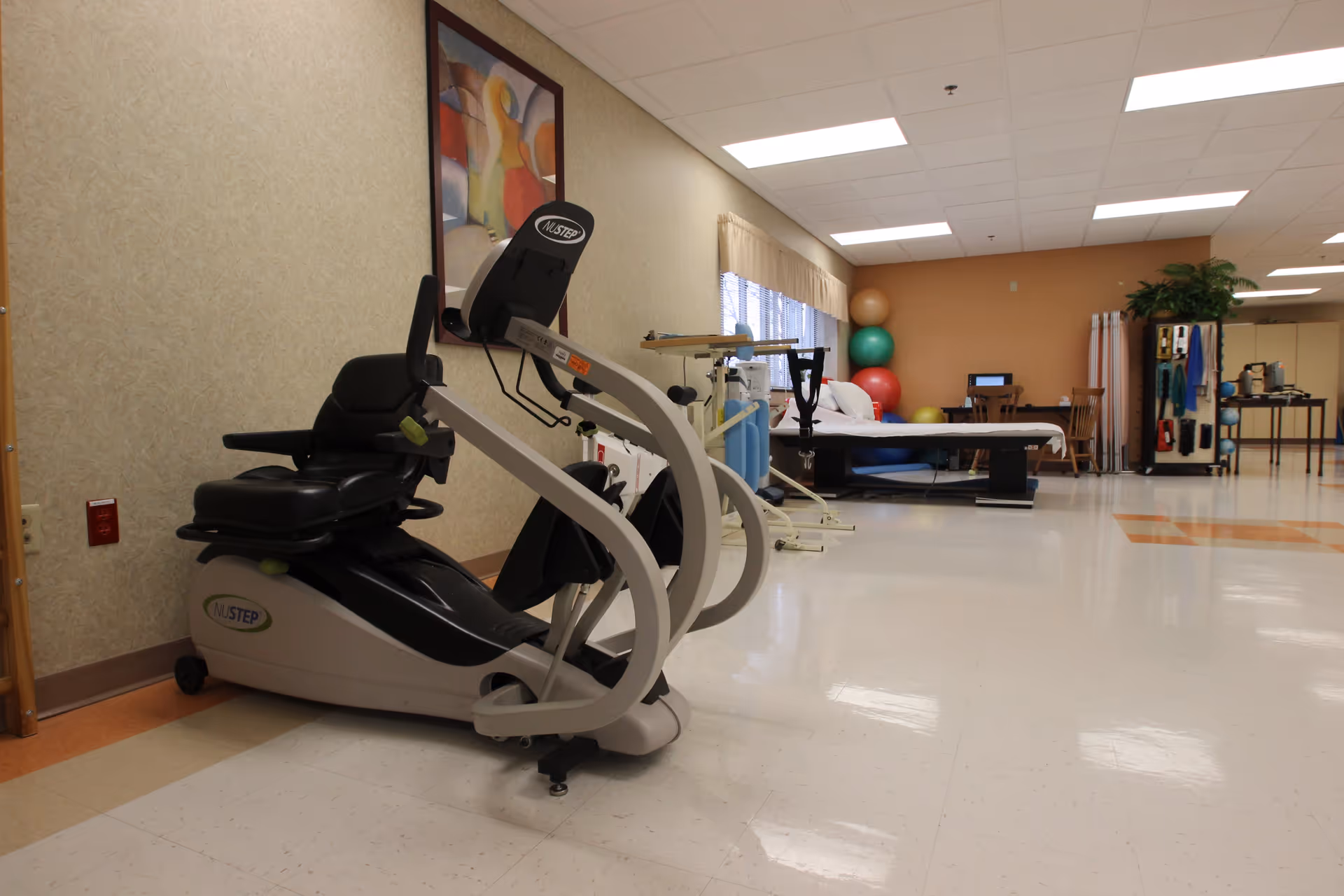 A physical therapy or rehabilitation room with exercise equipment including a NuStep recumbent cross trainer, therapy bed, exercise balls stacked in the corner, and various therapy tools and equipment along the walls. The room has beige walls, a tiled floor, and fluorescent ceiling lights.