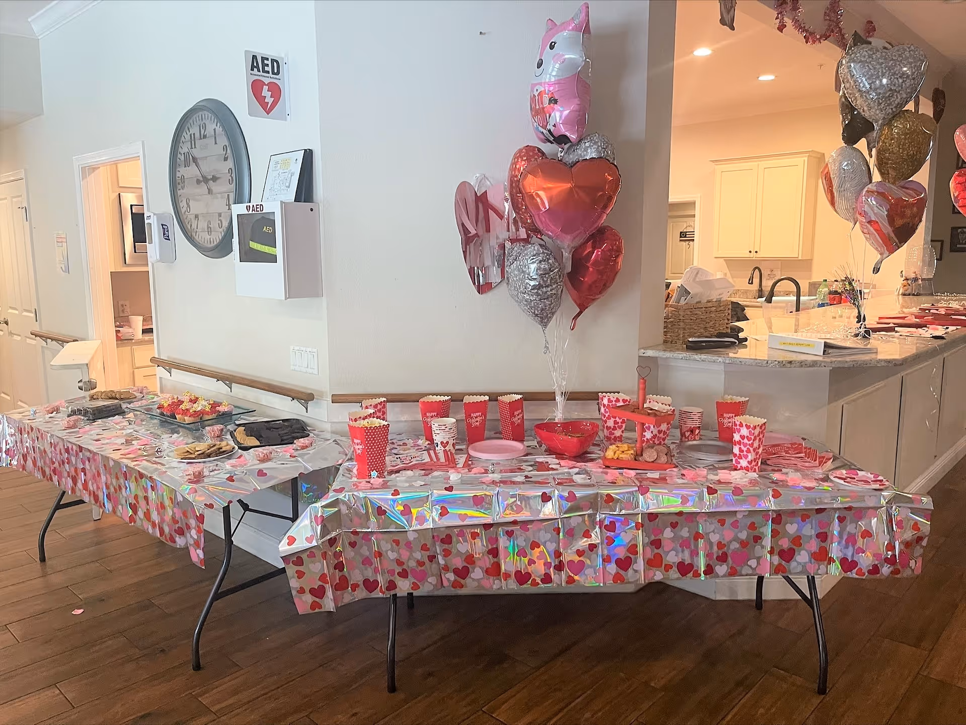 Two tables covered with heart-themed tablecloths and decorated with heart-shaped balloons and a unicorn balloon. The tables hold various snacks including popcorn, cookies, and cupcakes. The setting is an interior space with a clock and AED mounted on the wall, and a kitchen area visible in the background.