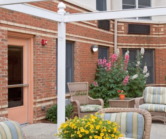 Outdoor patio area at The Pines of Mount Lebanon with wicker chairs and cushioned seating arranged around a small table. There are colorful flowers and plants, including yellow and pink blooms, against a brick building wall with windows and a door.