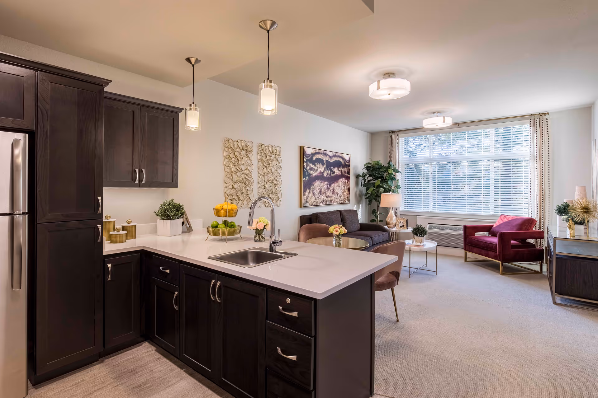 A modern open-concept living space featuring a kitchen with dark wood cabinets, a white countertop island with a sink, and stainless steel refrigerator. The living area includes a dark sofa, a red armchair, a coffee table, decorative plants, and large windows with blinds allowing natural light to fill the room.