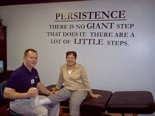 A healthcare worker is assisting an elderly woman by holding and stretching her leg while she sits on a padded examination table in a therapy or rehabilitation room. On the wall behind them is a motivational quote about persistence.