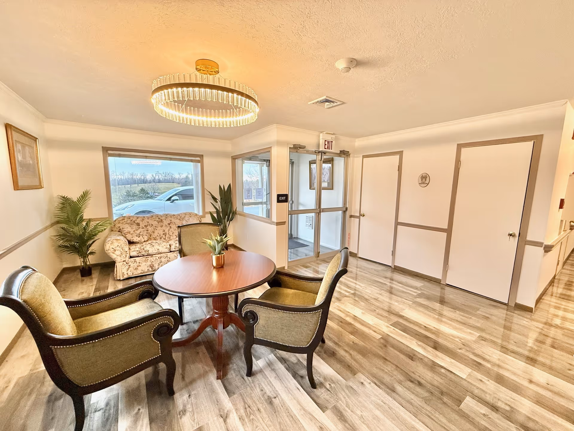 Small reception seating area with a round table and chairs, a floral loveseat, potted plants, and glass entry doors.
