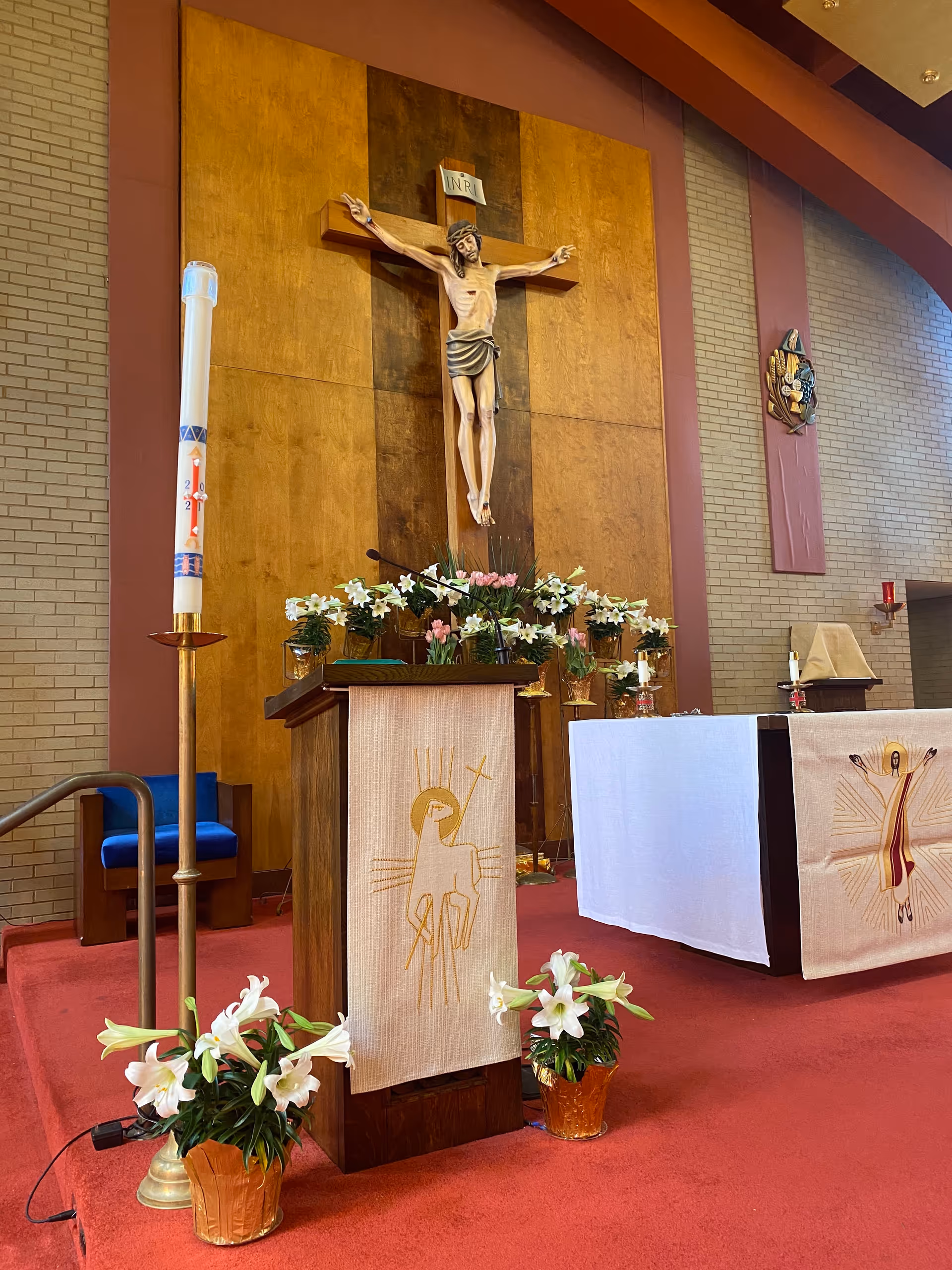 Interior view of a chapel with a large wooden crucifix mounted on a wood-paneled wall. Below the crucifix is a lectern draped with a cloth featuring a religious symbol, surrounded by white lilies in pots. To the right is an altar covered with white and beige cloths adorned with religious imagery. The floor is carpeted in red, and there is a tall candle on a stand to the left.