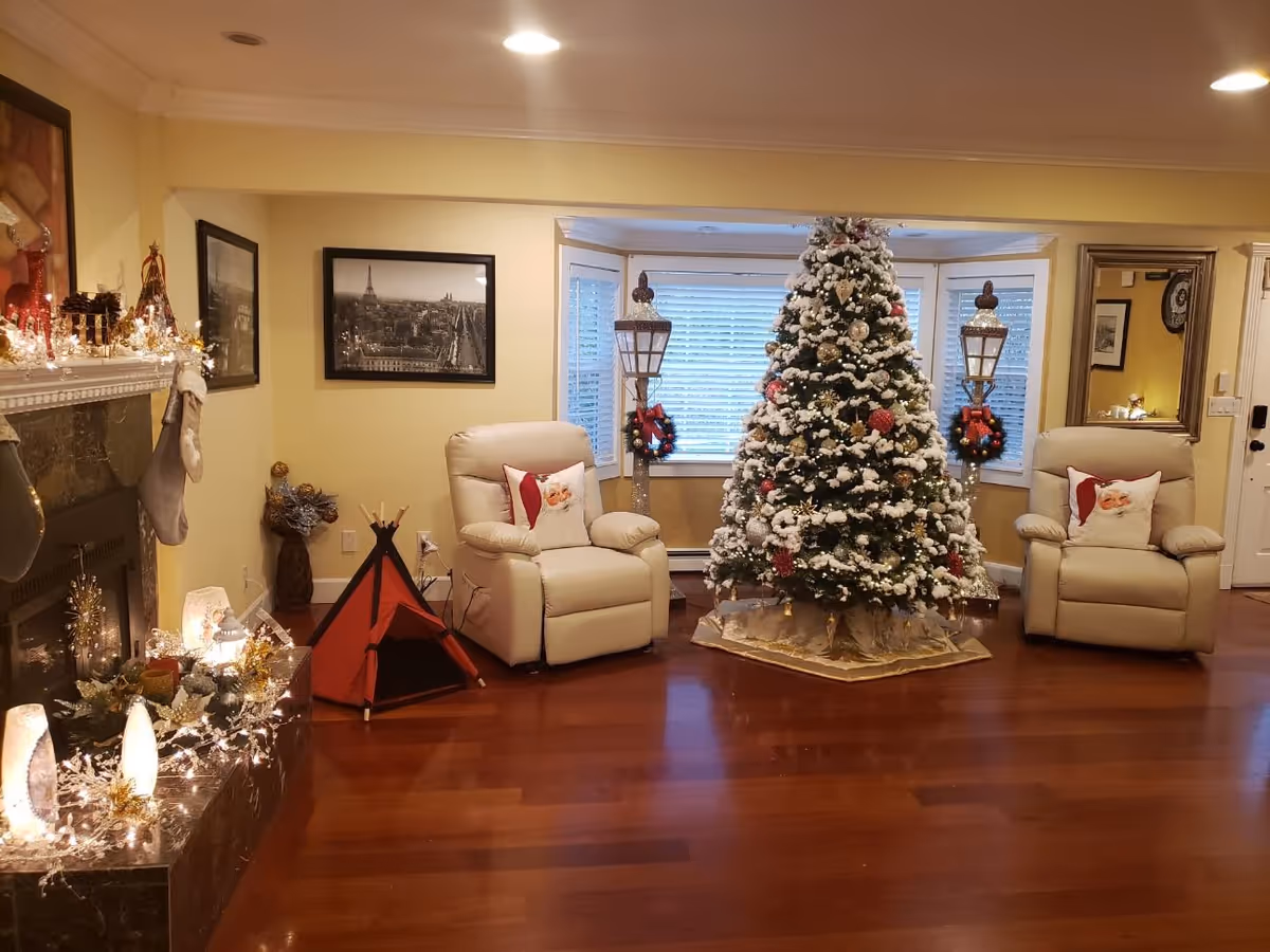 A cozy living room decorated for Christmas with a large, snow-covered Christmas tree adorned with ornaments and lights in front of a bay window. Two beige recliner chairs with Santa Claus themed pillows are placed on either side of the tree. The room features wooden flooring, a fireplace mantle decorated with Christmas lights, stockings, and festive ornaments. A small red and black play tent is positioned near one of the chairs. The walls are painted yellow and decorated with framed black and white photographs.