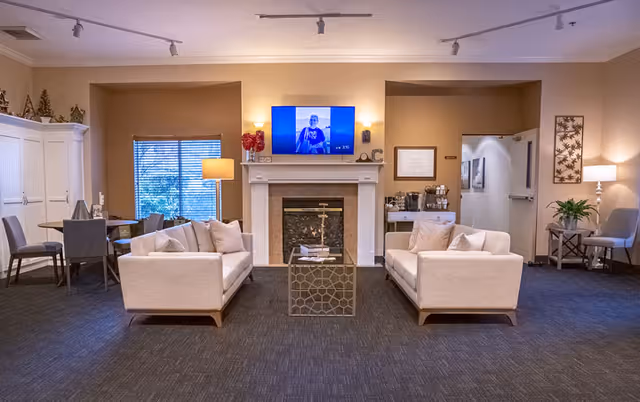 A cozy living room area in Cristwood Park featuring two beige sofas facing each other with a glass coffee table in between. A fireplace with a mounted flat-screen TV above it is centered on the back wall. To the left, there is a window with blinds and a floor lamp, and a small dining table with chairs. To the right, there is a side table with a lamp and a plant, and a hallway leading to other rooms. The room is softly lit with ceiling track lights and wall sconces.