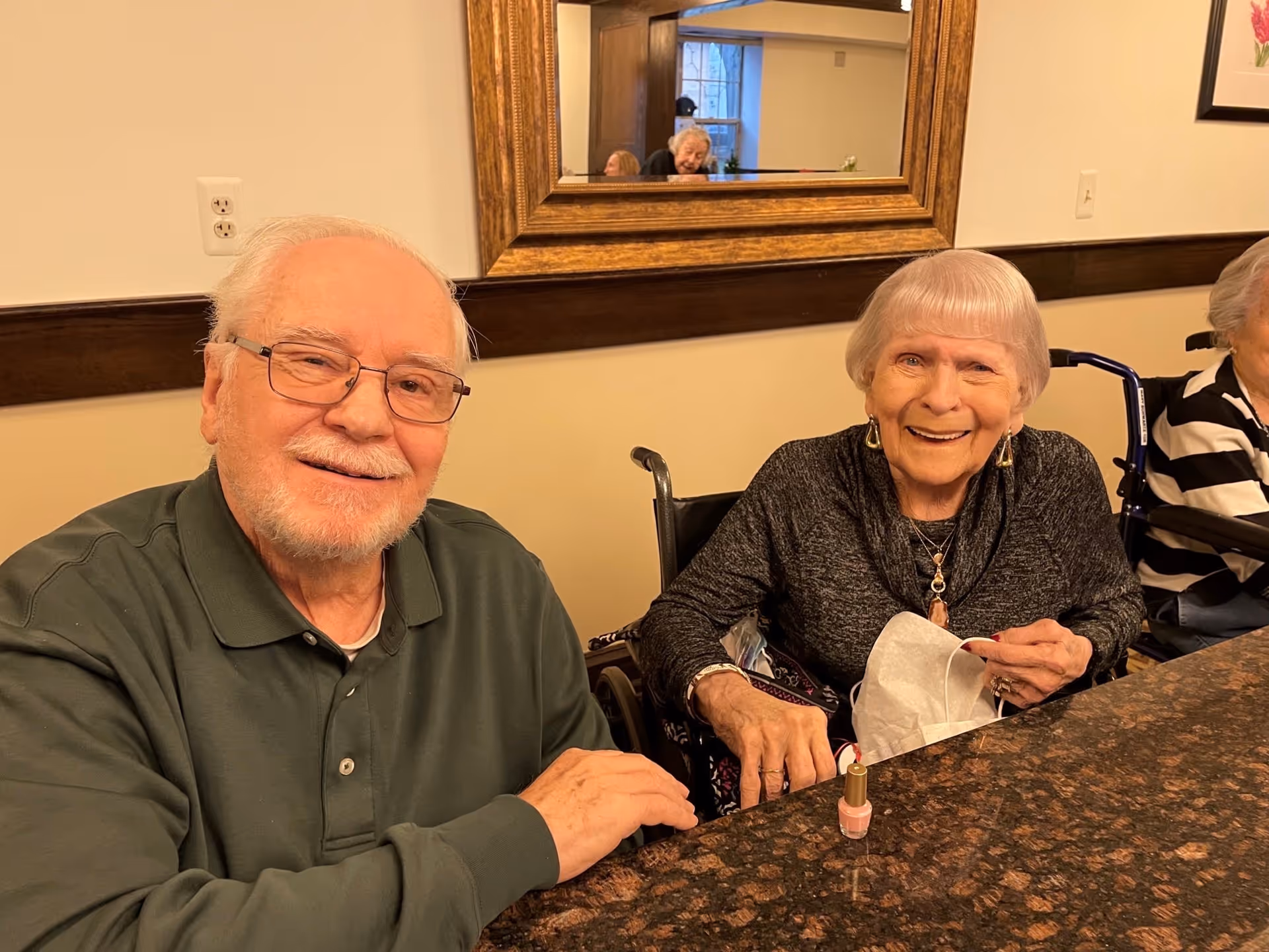 An elderly man and woman sitting at a granite table inside a senior living facility. The man is wearing glasses and a green polo shirt, smiling at the camera. The woman, also smiling, has short gray hair and is wearing a dark sweater and jewelry. There is a mirror on the wall behind them reflecting part of the room and other residents.