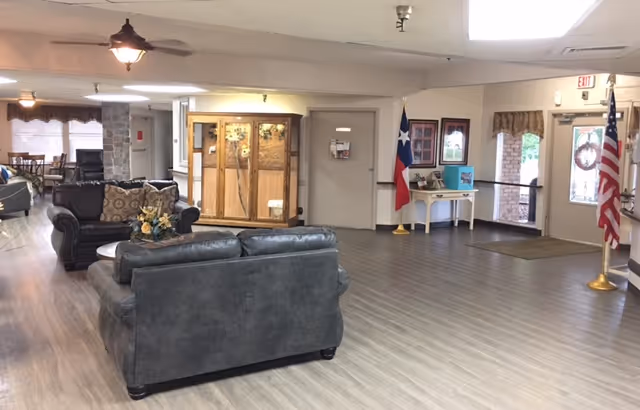 A spacious interior common area with gray wood flooring, two dark leather sofas with patterned cushions, a round coffee table with a floral arrangement, and a display cabinet against the wall. The room has a Texas state flag and an American flag near the entrance door, with framed pictures and a small table along the wall. The area is well-lit with ceiling lights and has large windows with curtains in the background.