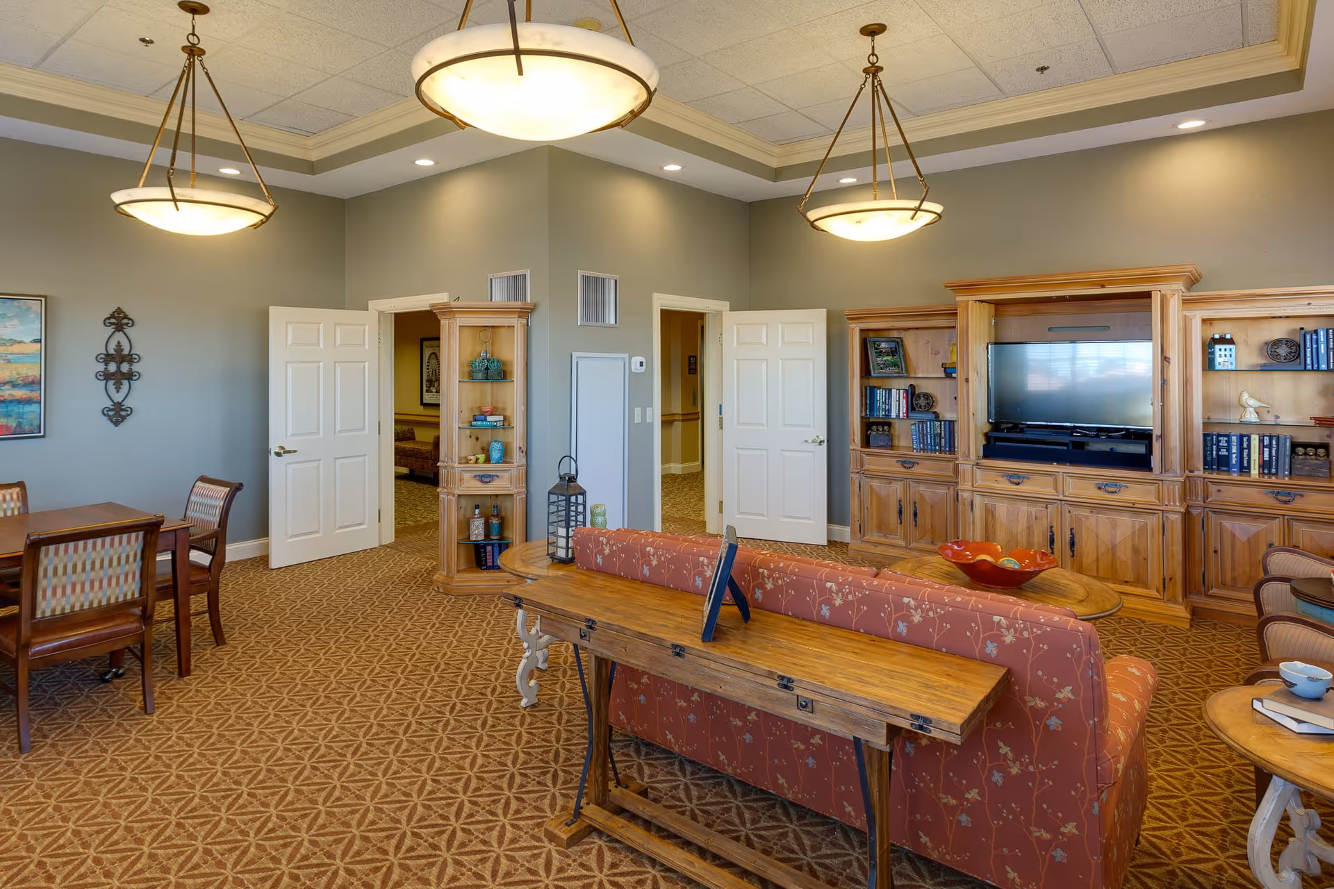 A cozy living room area in a senior living facility with a patterned carpet, a floral upholstered sofa, wooden furniture including a TV cabinet with a flat-screen TV, bookshelves, a wooden table behind the sofa, and a dining table with chairs. The walls are painted light gray with decorative wall art, and the ceiling has recessed lighting and two hanging light fixtures.