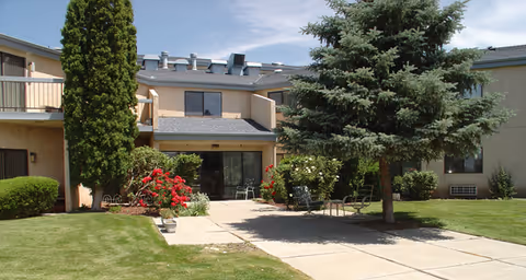Outdoor view of a senior living facility courtyard with a paved walkway, green grass, bushes, red flowers, and tall trees. The building is two stories with beige walls and several windows. There are a few chairs placed near the building entrance.