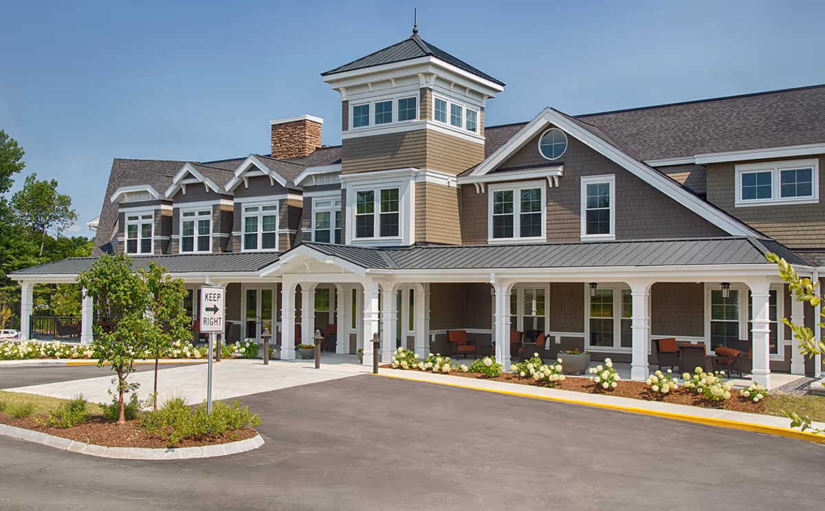 Front exterior of a two-story senior living facility with a covered porch entrance, tower feature, and landscaped driveway.