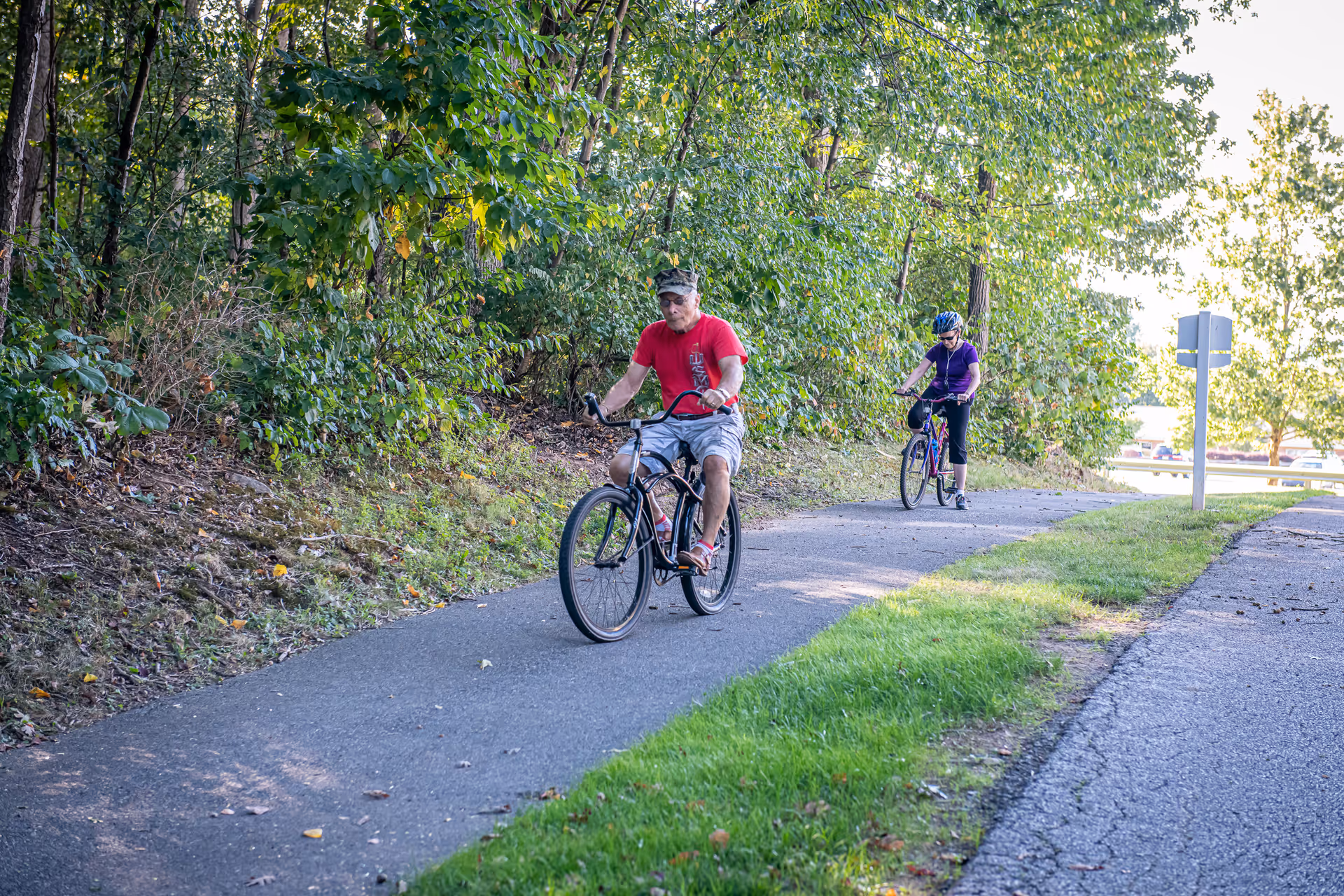 Two people riding bicycles on a paved path surrounded by green trees and grass on a sunny day.