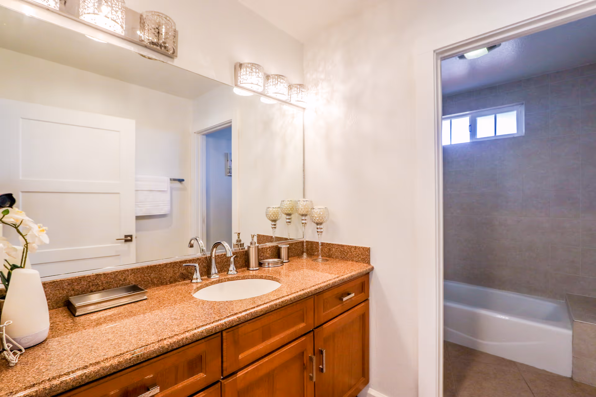 A bathroom vanity with a brown granite countertop, an oval sink, and silver faucet fixtures. Above the vanity is a large mirror with three decorative light fixtures mounted on the wall. On the countertop, there are decorative candle holders, a soap dispenser, and a small tray. To the right, there is an open doorway leading to a tiled bathtub and shower area with a small window above.