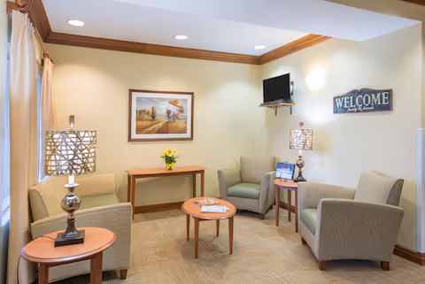 A cozy living room area with four beige armchairs arranged around a small round wooden coffee table. There are two side tables with decorative lamps, a small wooden console table with a vase of yellow flowers, a wall-mounted flat screen TV, and a framed painting on the wall. A 'WELCOME' sign is also displayed on the wall.