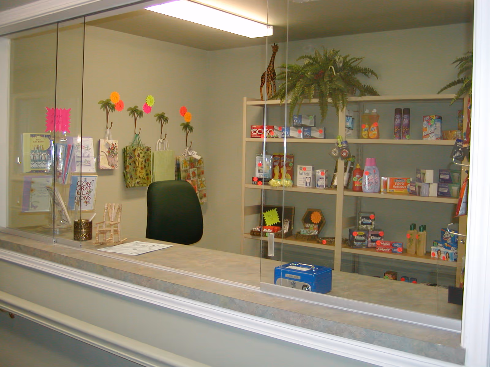A small retail or reception area with a glass partition. Behind the glass, shelves are stocked with various personal care products, toiletries, and small gift items. A black office chair is positioned behind the counter, and several gift bags hang on the wall. The area is well-lit with a fluorescent ceiling light.