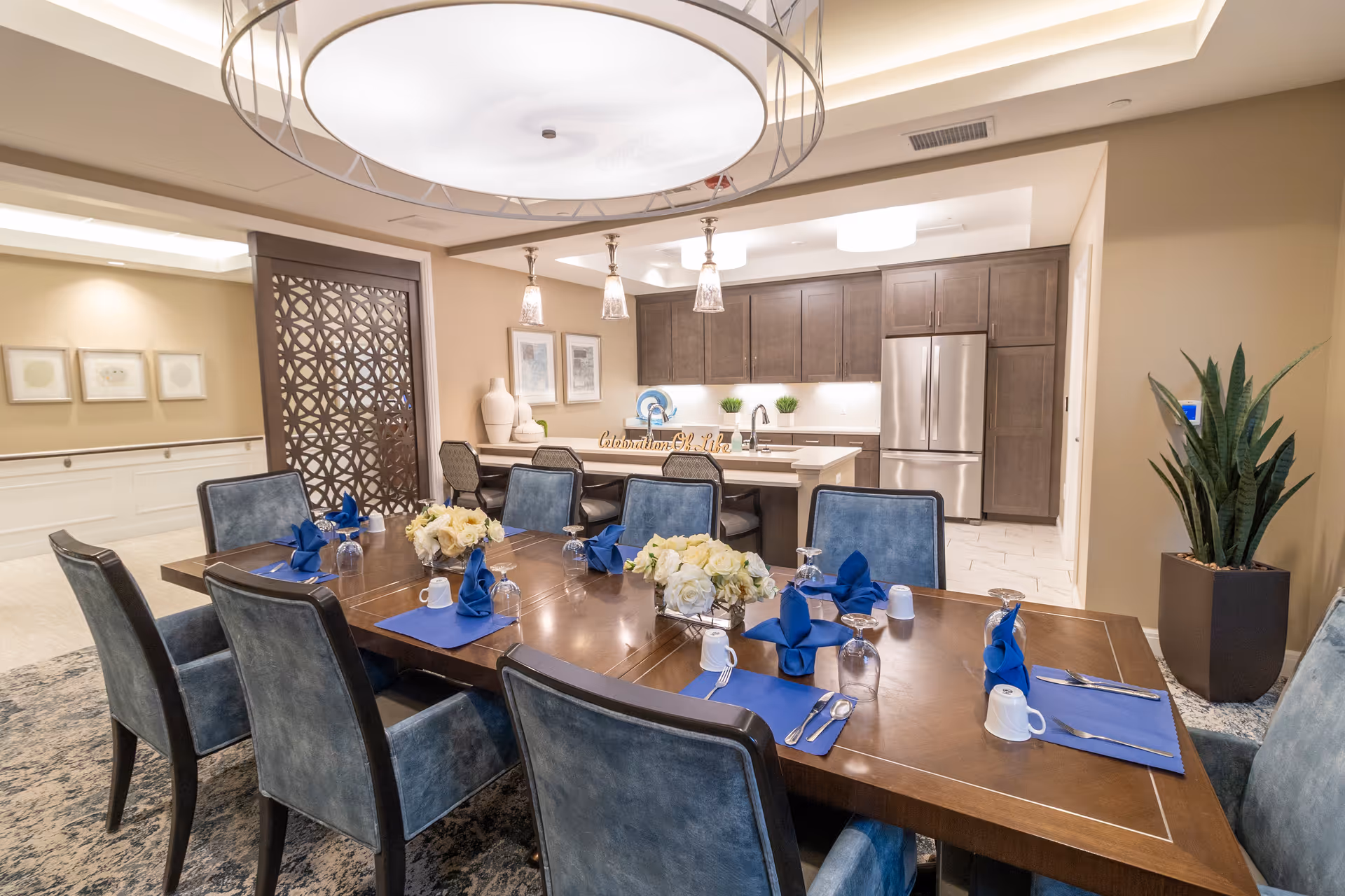 Elegant dining room with a large set table and blue chairs, open to a modern kitchen with dark cabinets in the background.