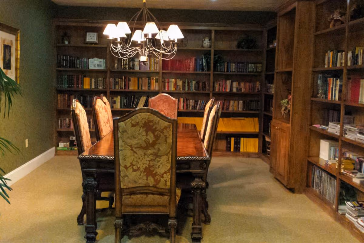 A cozy room with a long wooden dining table surrounded by six upholstered chairs. The walls are lined with wooden bookshelves filled with books and some decorative items. A chandelier with multiple lampshades hangs above the table, providing warm lighting.