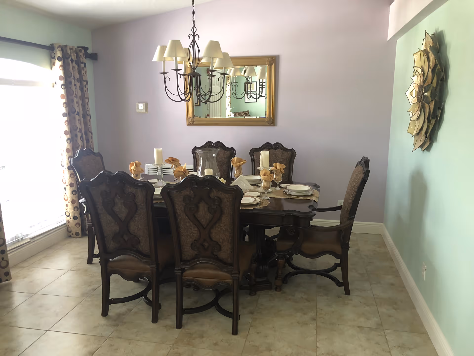 A dining room with a dark wooden dining table set for six people. The table has ornate chairs with upholstered backs and seats. There are folded napkins, plates, and candles on the table. A chandelier with multiple lampshades hangs above the table. A large mirror with a gold frame is mounted on the light purple wall behind the table. The floor is tiled, and there are patterned curtains on the window to the left. A decorative golden wall art piece is on the light blue wall to the right.