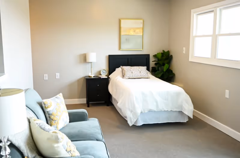Small, bright bedroom with a single bed against a gray wall, a nightstand, a sofa, and a window.