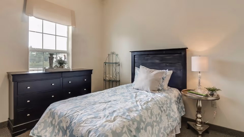 A bedroom with a single bed featuring a black wooden headboard and light blue floral bedding. Next to the bed is a round metallic side table with a lamp, a small plant, and some books. Across from the bed is a black dresser with multiple drawers, and a window above it with a beige valance. The walls are light-colored, and there is a decorative metal shelf in the corner.