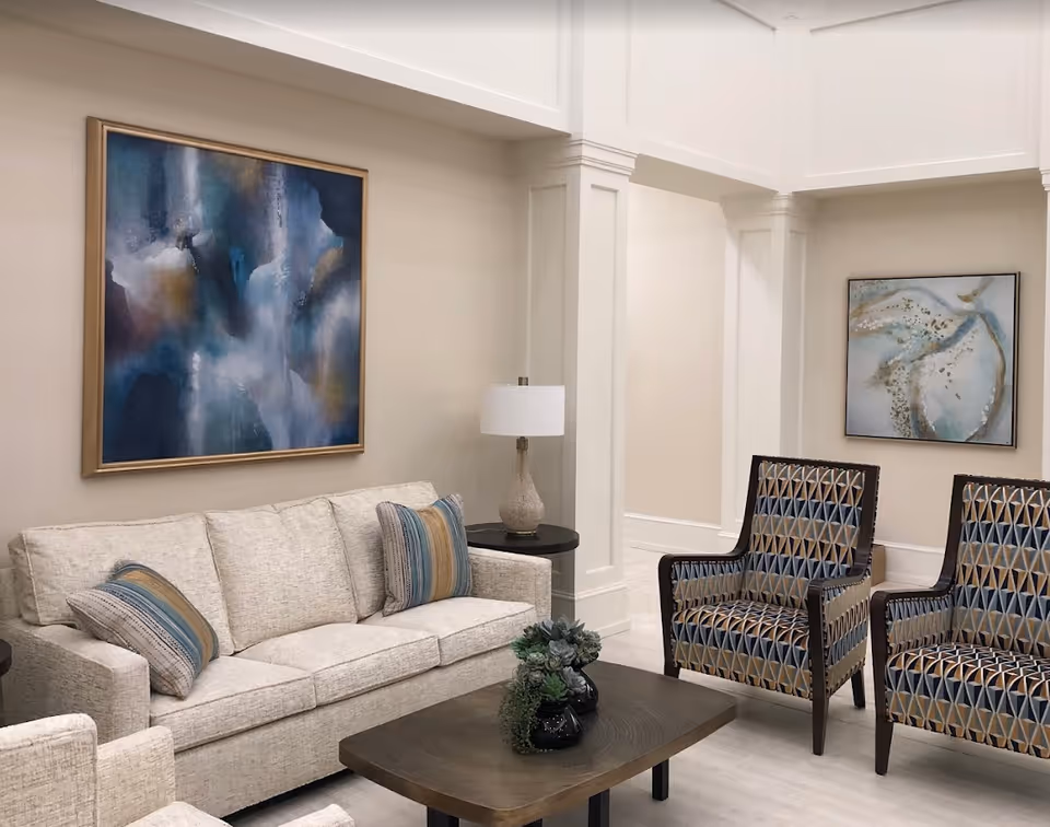 Well-lit seating area with a beige sofa, patterned armchairs, a coffee table, and abstract wall art on the walls.