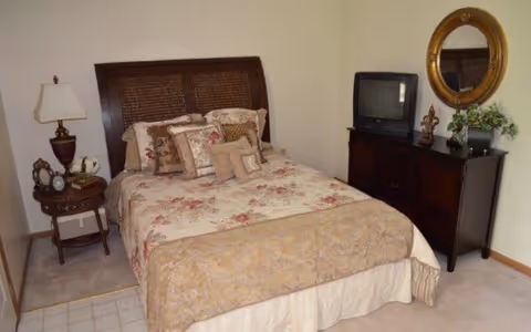 A bedroom with a bed featuring a floral patterned bedspread and multiple decorative pillows. To the left of the bed is a small round wooden side table with a lamp, clock, and other small items. On the right side of the room is a dark wooden dresser with a small TV on top, a decorative statue, a plant, and a round gold-framed mirror hanging on the wall above.