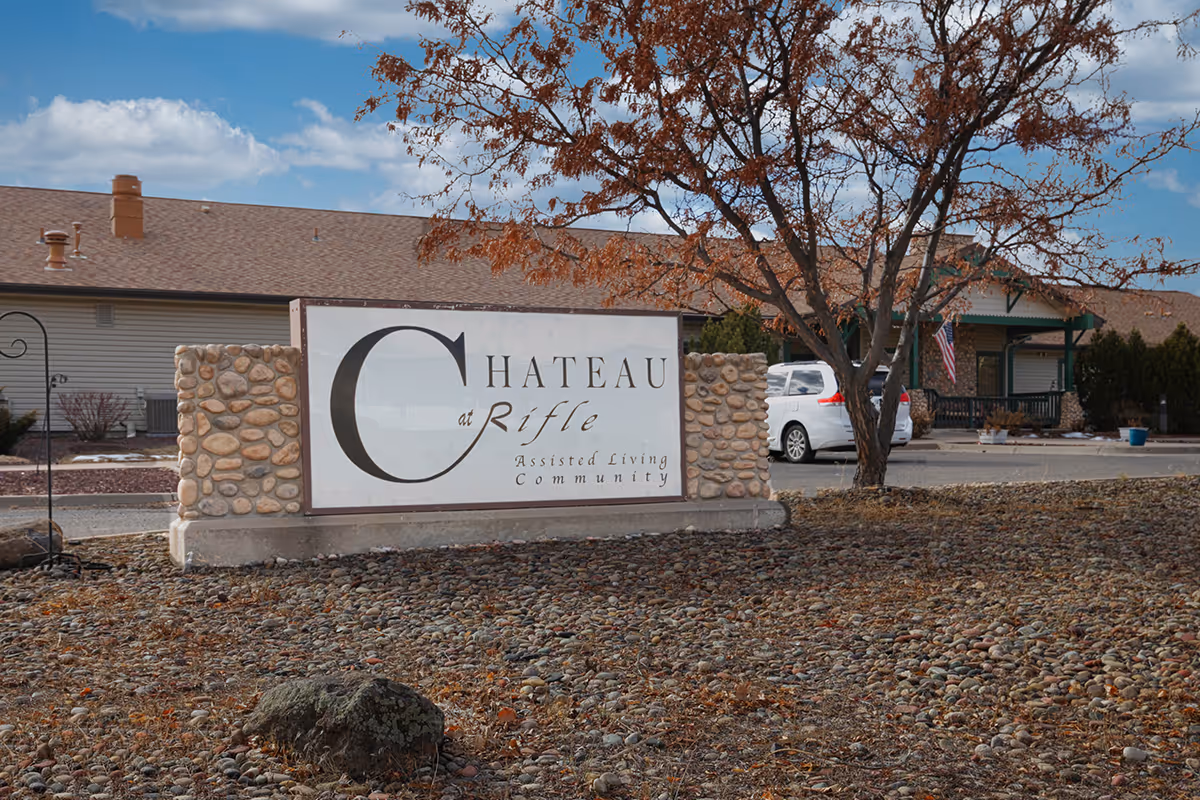 Outdoor view of the Chateau at Rifle assisted living community sign with a stone base and a building in the background under a partly cloudy sky. There is a tree with brown leaves and a white vehicle parked near the building.