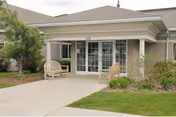 Front entrance of a single-story care facility with glass double doors, a covered porch, two wicker chairs, and a landscaped walkway.